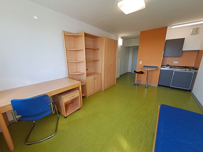 Ein helles, modernes Zimmer mit grünem Bodenbelag. Es gibt einen Schreibtisch mit blauem Stuhl, ein Regal, einen Kleiderschrank und eine Küchenecke mit schwarzem Kühlschrank und grauer Arbeitsplatte. Ein hoher Hocker steht an einem kleinen Tisch in der Nähe der Küche.
