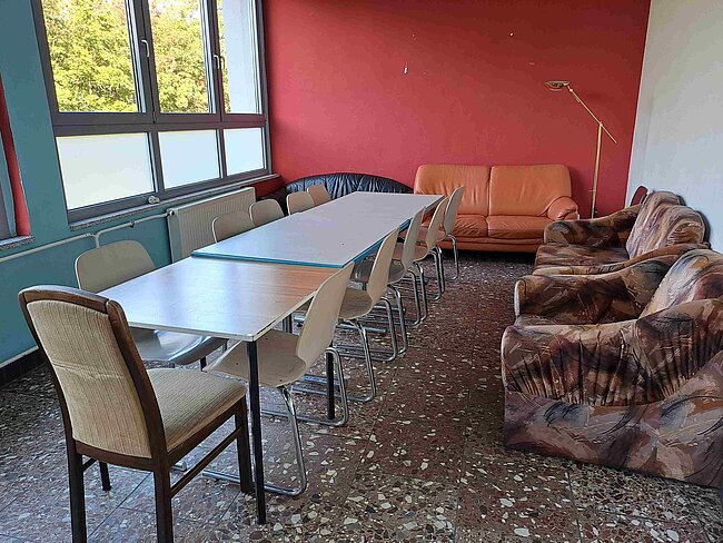 Ein Raum mit einem langen Tisch und mehreren Stühlen, einem abgenutzten braunen Sofa und einem weiteren orangefarbenen Sofa. Die Wände sind rot, und es gibt große Fenster an einer Seite. Der Boden ist mit Fliesen bedeckt, und der Raum wirkt etwas heruntergekommen.