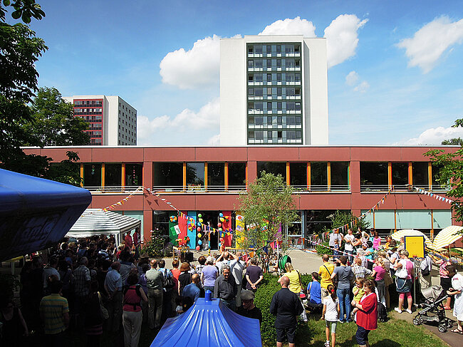Eine belebte Veranstaltung vor einem roten Gebäude mit mehreren Stockwerken. Viele Menschen, darunter Kinder und Erwachsene, sind versammelt. Bunte Zelte und Stände sind aufgestellt, und die Atmosphäre wirkt festlich. Der Himmel ist blau mit einigen Wolken.