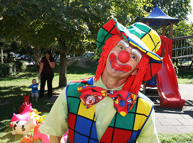 Ein Clown mit roter Perücke, bunter Kleidung und einer großen Mütze steht in einem Park. Er trägt eine rote Nase und lächelt. Im Hintergrund sind ein Kind, eine Erwachsene und eine rote Rutsche zu sehen. Der Clown hält einige Spielzeuge in der Hand.