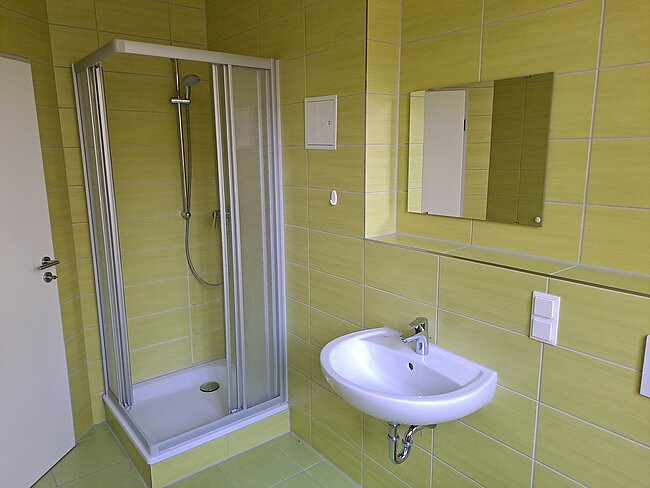 Ein modernes Badezimmer mit gelben Fliesen. Es gibt eine Duschkabine mit Glaswänden und einem Duschkopf, ein Waschbecken mit einem Wasserhahn und einen großen Spiegel über dem Waschbecken. An der Wand befindet sich ein Lichtschalter und ein Thermostat.