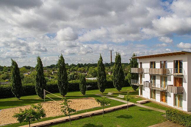 Ein modernes Gebäude mit mehreren Balkonen, umgeben von einem gepflegten Garten mit Bäumen und einem Sandplatz, wahrscheinlich für Sport oder Freizeitaktivitäten. Der Himmel ist teilweise bewölkt.