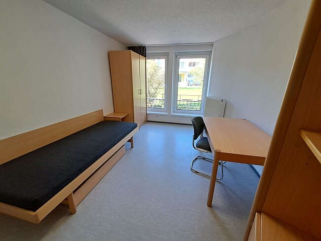Ein einfaches Zimmer mit einem ausziehbaren Bett, einem Schreibtisch mit Stuhl und einem Kleiderschrank. Das Zimmer hat ein Fenster mit Blick auf einen grünen Außenbereich. Die Wände sind weiß und der Boden ist mit Teppich ausgelegt.