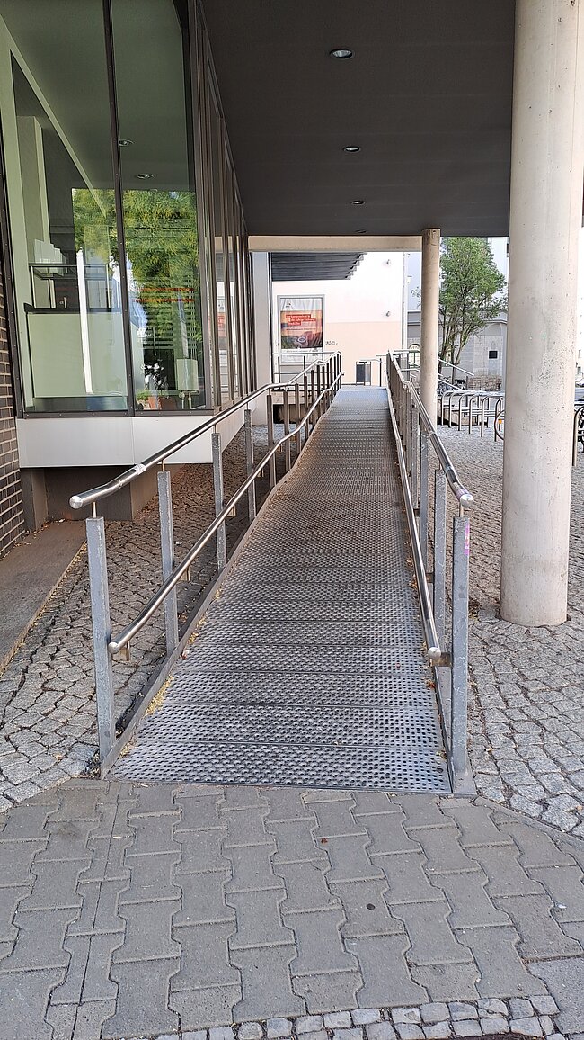 Ein barrierefreier Weg mit Metallgeländern führt zu einem Gebäude. Der Boden besteht aus Pflastersteinen und einem taktilen Leitsystem für Sehbehinderte. Im Hintergrund sind Bäume und ein Teil des Gebäudes mit Glasfronten zu sehen.