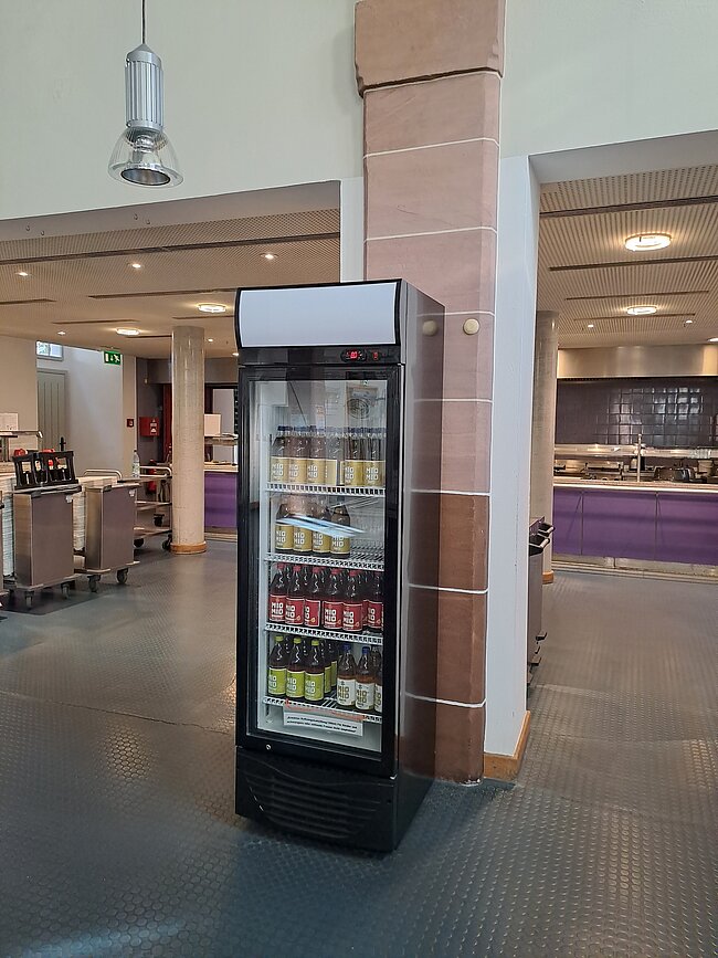 Ein Kühlregal mit verschiedenen Getränken steht in einem Raum mit Fliesenboden und Säulen. Im Hintergrund sind Tische und Stühle sowie ein Tresenbereich zu sehen. Der Raum wirkt wie eine Cafeteria oder ein Aufenthaltsbereich.