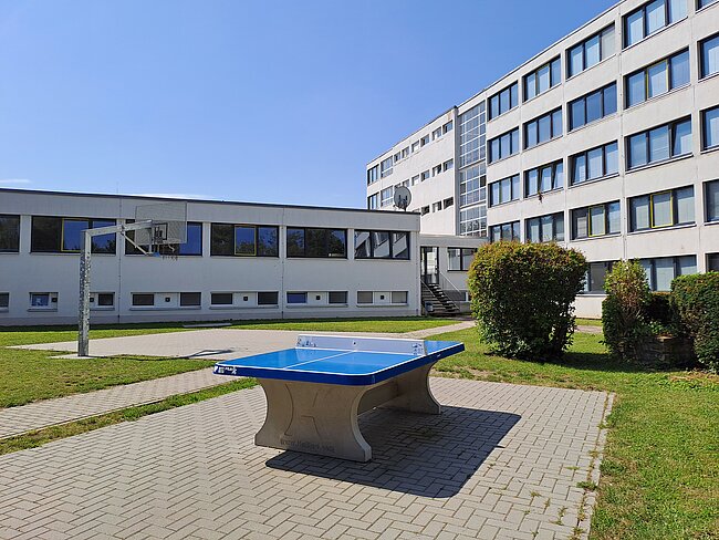 Ein blaues Tischtennis-Tisch steht auf einem gepflasterten Bereich vor einem modernen Gebäude mit vielen Fenstern. Im Hintergrund ist ein Rasenbereich mit Büschen zu sehen.