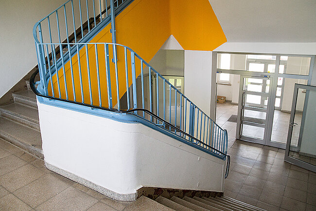 Treppenhaus Wohnheim Magdeburger Straße, Bernburg; Foto: Annegret Höhne Ein Treppenhaus mit einer weißen und blauen Wendeltreppe, die zu einer Tür mit Glasfenstern führt. Die Wände sind teilweise orange gestrichen. Der Boden besteht aus grauen Fliesen. Die Treppe hat ein blaues Geländer.