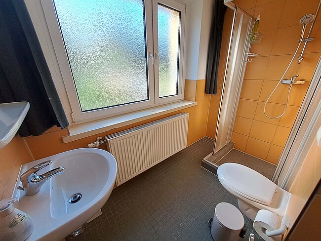 Ein modernes Badezimmer mit einem Waschbecken, einer Toilette und einer Duschkabine. Es gibt ein Fenster mit Milchglas und einen Heizkörper darunter. Die Wände sind in einem warmen Orangeton gefliest, und der Boden hat graue Fliesen.