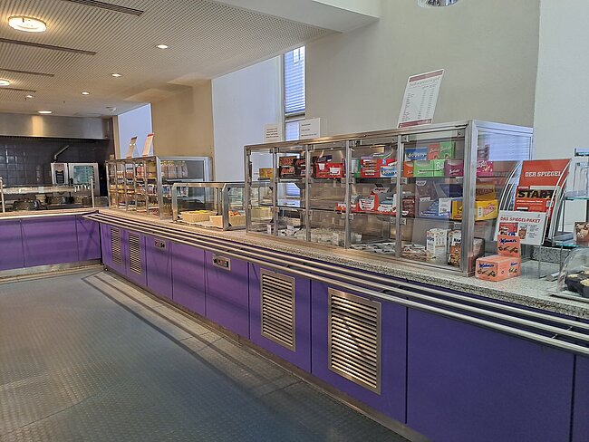 Ein Selbstbedienungsbereich in einem Café oder einer Kantine mit einer purpurfarbenen Theke. Auf der Theke stehen verschiedene Snacks, Getränke und Kaffeemaschinen. Im Hintergrund sind Regale mit weiteren Produkten zu sehen. Der Boden ist mit Fliesen bedeckt.
