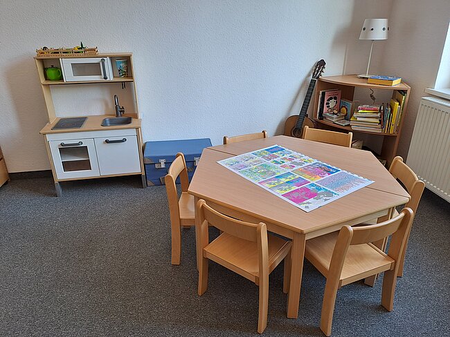 Ein Kinderzimmer mit einem kleinen Tisch und vier Stühlen. Auf dem Tisch liegt ein buntes Puzzle. Im Raum befindet sich eine Spielküche mit verschiedenen Kochutensilien. Ein Regal mit Büchern und ein Kinderwagen sind ebenfalls vorhanden. Der Boden ist mit Teppich ausgelegt.