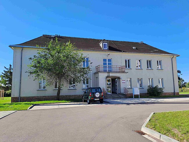 Ein zweistöckiges Gebäude mit weißer Fassade und einem schwarzen Auto davor. Das Gebäude hat mehrere Fenster und eine kleine Überdachung über dem Eingang. Vor dem Haus steht ein Baum, und der Himmel ist klar und blau.
