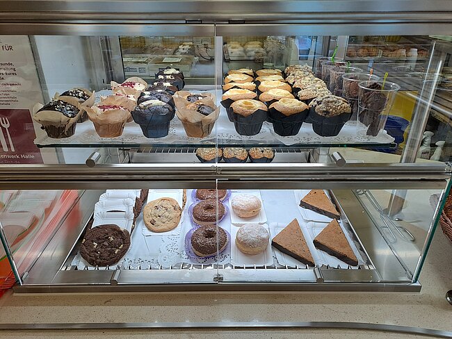Ein Bild eines Bäckereiregal mit verschiedenen Backwaren. Oben befinden sich Muffins mit verschiedenen Belägen wie Schokolade und Früchten. Unten sind Donuts, Kuchenstücke und andere Süßigkeiten. Die Auslage ist gut beleuchtet und die Backwaren sind ansprechend präsentiert.