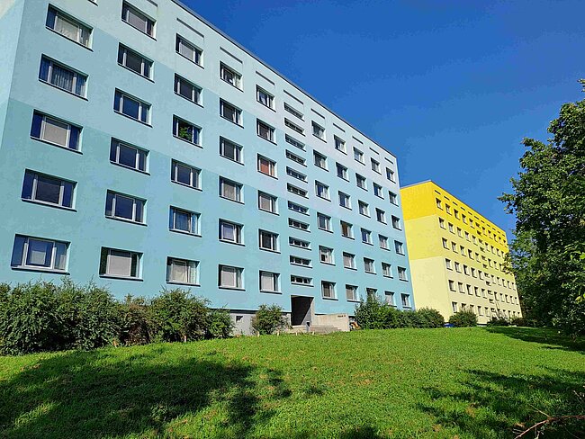 Ein mehrstöckiges Gebäude mit blauer und gelber Fassade, umgeben von grünem Gras und Büschen. Das Gebäude hat viele Fenster und wirkt wie ein Wohn- oder Bürokomplex. Der Himmel ist klar und blau.