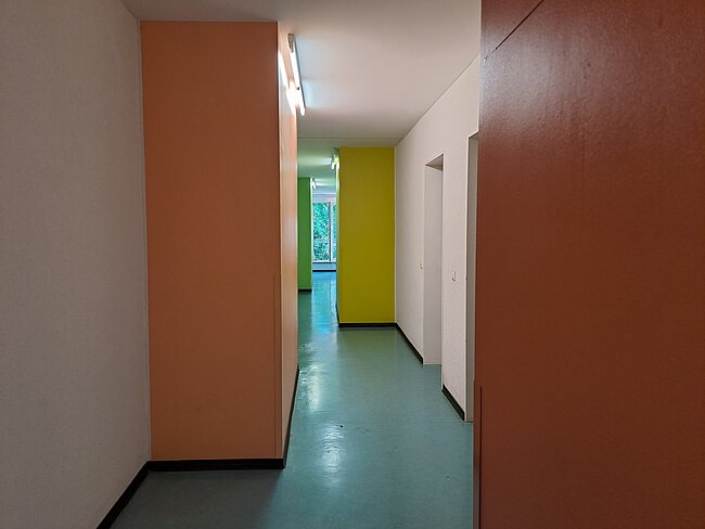 Ein langer Flur mit orangefarbenen Wänden auf der linken und rechten Seite. Der Boden ist grünlich-blau. Am Ende des Flurs befindet sich ein Raum mit Tageslicht. Es gibt eine Tür auf der rechten Seite des Flurs.