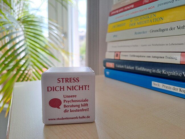 Würfel mit der Aufschrift „STRESS DICH NICHT!“ und Hinweis auf kostenfreie psychosoziale Beratung des Studentenwerks Halle mit Website-Angabe. Im Hintergrund Bücher zum Thema Meditation und Selbsthilfe.