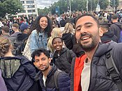 Eine Gruppe von fünf Personen lächelt in die Kamera. Sie stehen bei einer Menschenansammlung im Freien, möglicherweise bei einer Veranstaltung oder Demonstration. Die Menschen im Hintergrund sind ebenfalls versammelt und scheinen aktiv zu sein.