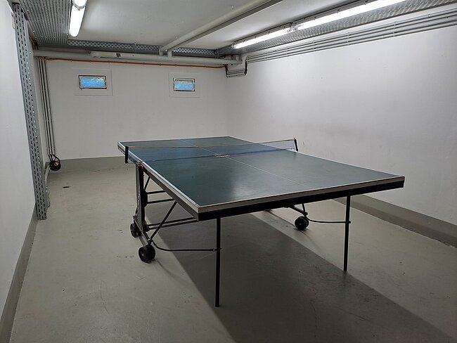 Tischtennisplatte Wohnheim Brandbergweg, Foto: S. Loreck Ein Tischtennistisch steht in einem hellen, kahlen Raum mit Betonboden. Der Tisch hat eine graue Oberfläche und ist auf Rollen platziert. Die Wände sind weiß, und es gibt zwei kleine Fenster oder Lüftungsschlitze. Deckenleuchten sorgen für ausreichend Licht.