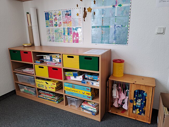 Ein Regal mit bunten Fächern und Aufbewahrungsboxen, gefüllt mit Spielzeug, Büchern und Lernmaterialien wie Bausteine, Puzzles und Stifte. Daneben ein kleiner Schrank mit Spielkleidung und Accessoires.