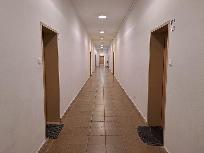 Ein langer, beleuchteter Flur mit weißen Wänden und hellen Fliesen auf dem Boden. Auf beiden Seiten befinden sich Türen mit Nummern, darunter Fußabstreifer. Das Licht kommt von der Decke und der Flur wirkt sauber und ordentlich.
