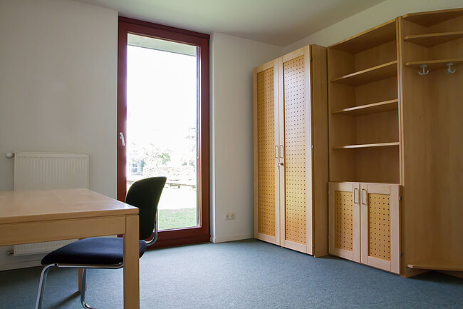 Ein helles, minimalistisch eingerichtetes Zimmer mit einem Schreibtisch, einem Stuhl, einem großen Schrank und einem Regal. Ein Fenster mit Blick auf eine Terrasse oder einen Balkon lässt viel Licht herein. Der Boden ist mit blauem Teppich ausgelegt.