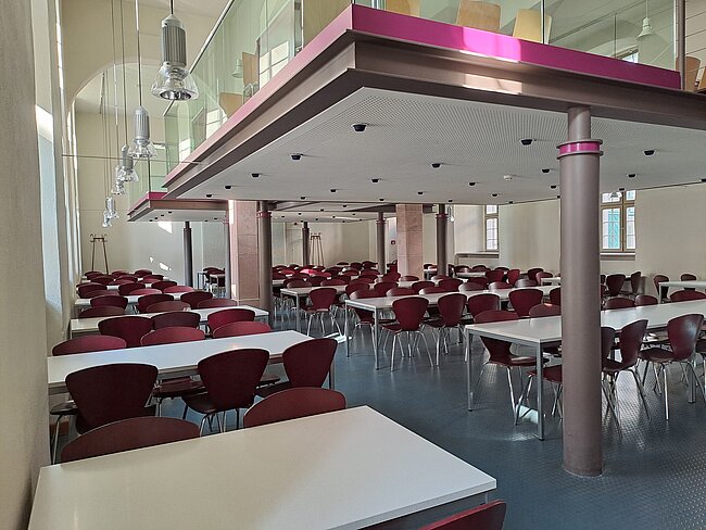 Ein großer, leerer Speisesaal mit vielen Tischen und Stühlen. Die Tische sind weiß und die Stühle sind rot. Der Raum hat große Fenster und eine moderne Decke mit pinken Akzenten. Es gibt auch einige Säulen und moderne Lampen an der Decke.