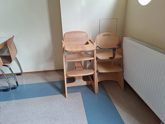 Hochstühle für die Kleinsten "Zwei Kinderhochstühle aus Holz stehen nebeneinander in einem Raum mit blauem und grauem Boden. Im Hintergrund befindet sich ein Radiator und ein Teil eines Schreibtischstuhls. Die Wände sind hell gestrichen."