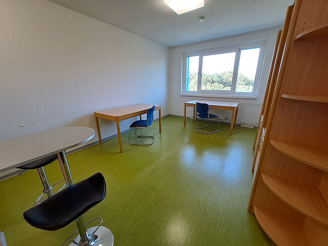 Ein helles Zimmer mit grünem Boden. Es gibt zwei Schreibtische mit Stühlen, einen runden Tisch mit einem Hocker und ein Regal. Das Zimmer hat ein großes Fenster mit Blick auf Bäume und eine Deckenlampe. Die Wände sind weiß und der Raum wirkt sauber und aufgeräumt.
