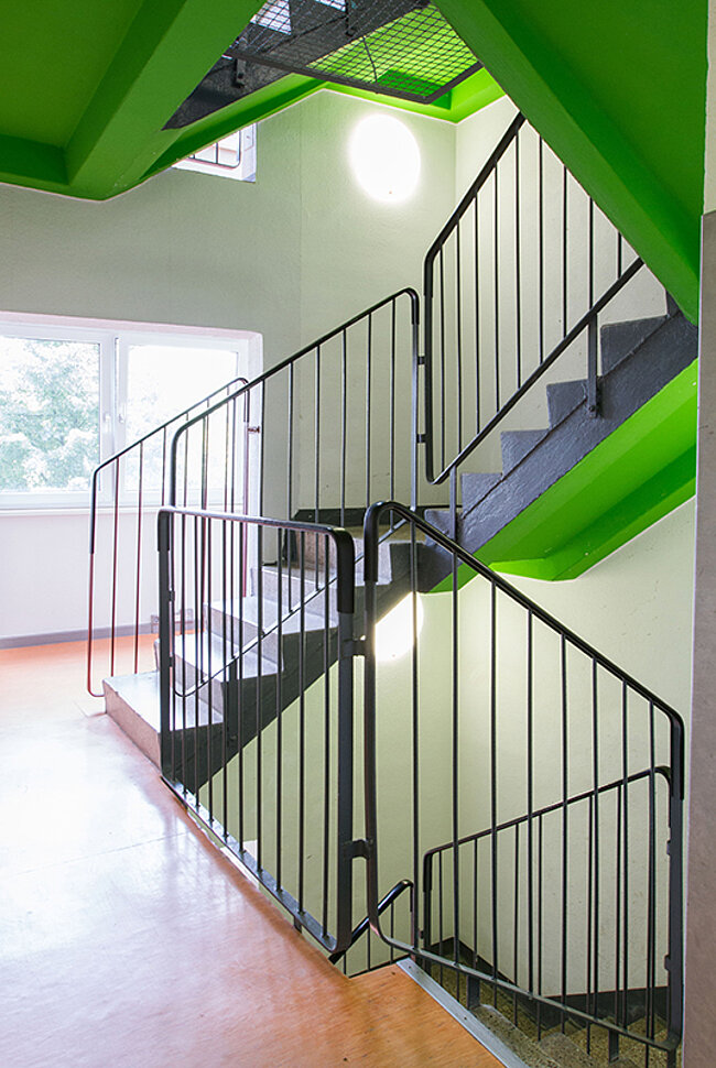 Eine Treppe mit grünen Wänden und einem schwarzen Geländer. Die Treppe führt nach oben und hat eine Wendelstruktur. Im Hintergrund befindet sich ein Raum mit Fenstern und Tageslicht. Die Decke ist ebenfalls grün mit einer Lichtquelle in der Mitte.
