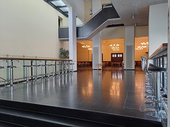 lichtdurchfluteter Gang im 1. OG mit Barhockern Ein moderner Innenraum mit glänzendem Boden, einer Glasbalustrade und mehreren Stühlen. Im Hintergrund sind Tische und Stühle in einem Speisebereich zu sehen. Deckenleuchten hängen von der hohen Decke herab, und große Fenster lassen natürliches Licht herein.