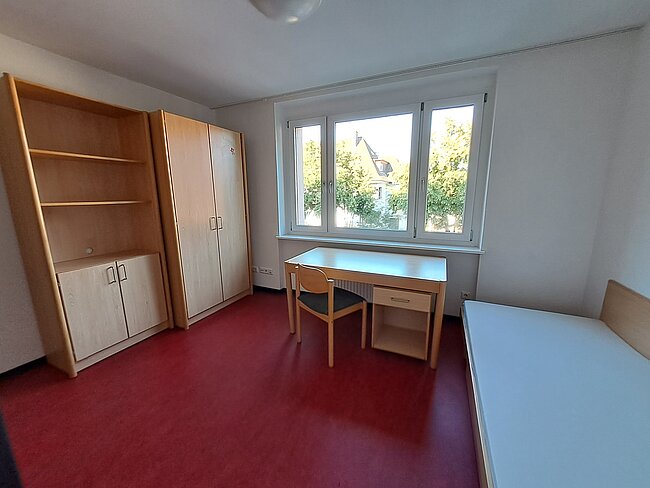 Ein einfaches, helles Zimmer mit rotem Teppichboden. Es enthält einen Schreibtisch mit Stuhl, ein Bett, einen großen Schrank und ein Regal. Die Fenster bieten Blick auf Bäume und einen Kran. Das Zimmer wirkt sauber und funktional eingerichtet.
