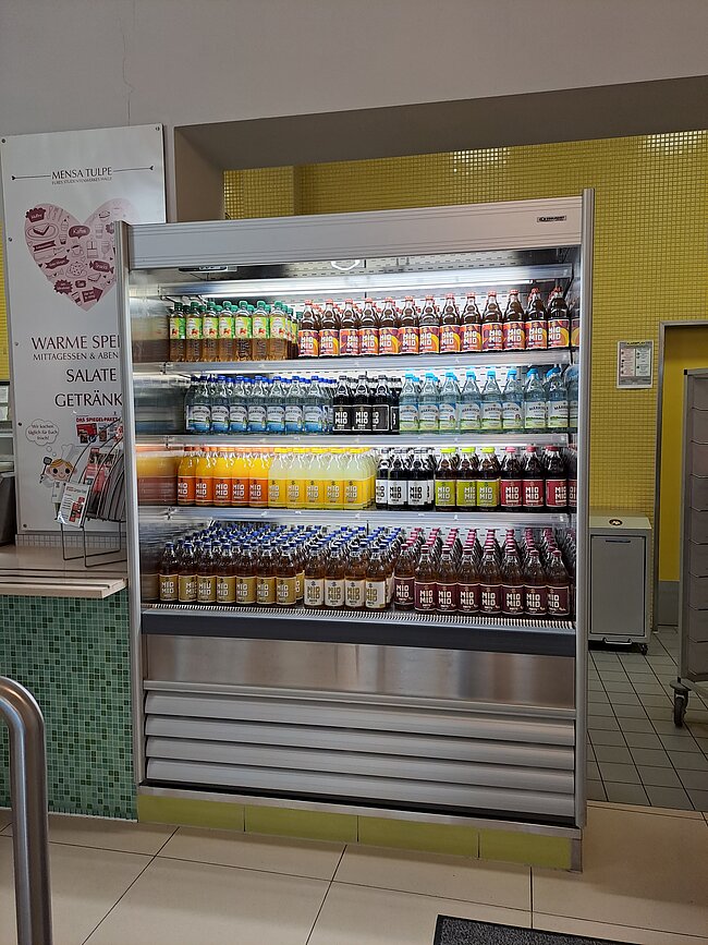 Ein Kühlregal in einem Supermarkt mit verschiedenen Getränken und Snacks. Die Regale sind gut sortiert und enthalten Flaschen, Dosen und verpackte Lebensmittel. Auf einem Schild steht "Warme Speisen, Mittagessen & Snacks, Salate, Getränke".