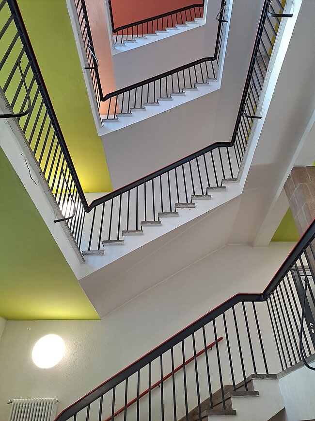 "Ein mehrstöckiges Treppenhaus mit mehreren Ebenen, das durch eine zentrale Öffnung verbunden ist. Die Wände sind in verschiedenen Farben gestrichen: gelb, grün und weiß. Die Treppen und Geländer sind aus Holz und Metall. Das Licht kommt von einer Deckenlampe."