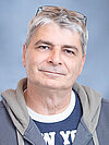 Ein älterer Mann mit grauen Haaren und Brille lächelt leicht. Er trägt ein T-Shirt mit der Aufschrift "NEW YORK" und eine graue Jacke. Der Hintergrund ist blau.
