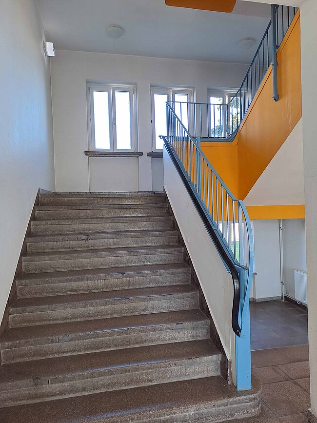 Ein Treppenhaus mit einer breiten Treppe, die nach oben führt. Die Wände sind weiß gestrichen, und es gibt ein orangefarbenes Geländer. Oben befindet sich ein Fenster, das Tageslicht hereinlässt. Die Stufen sind aus grauem Stein.