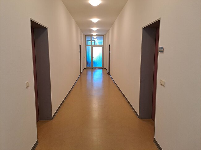 Ein langer, schmaler Flur mit weiß gestrichenen Wänden und einem hellen Boden. An beiden Seiten befinden sich Türen, und am Ende des Flurs ist eine Glastür mit Tageslicht zu sehen. Deckenleuchten erhellen den Gang.