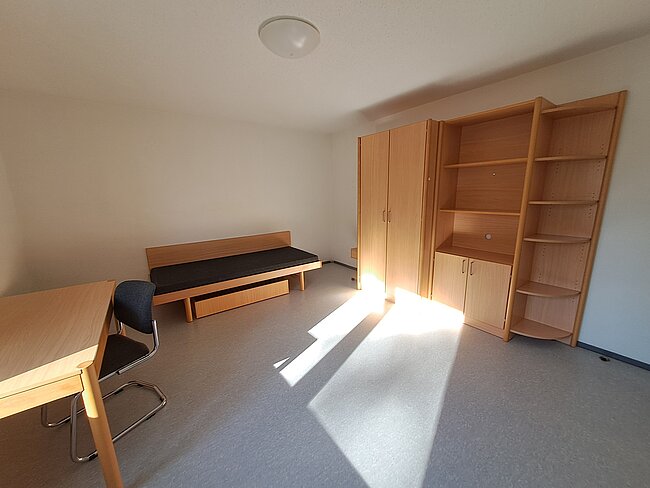 Ein einfaches Zimmer mit einem Bett, einem Schreibtisch mit Stuhl und einem Schrank. Das Zimmer hat helle Wände und einen grauen Teppichboden. Sonnenlicht fällt durch die Tür herein und wirft Schatten auf den Boden.
