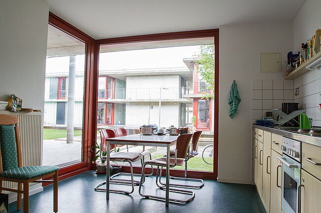 Ein helles Wohnzimmer mit Küche, das durch eine große Fensterfront mit Blick auf Wohngebäude beleuchtet wird. Im Raum befinden sich ein Esstisch mit vier Stühlen, eine Küchenzeile und ein bequemer Sessel. Die Einrichtung ist modern und funktional.