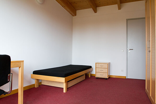 Ein einfaches Zimmer mit einem schmalen Bett, einem kleinen Schreibtisch mit Stuhl und einem dreistöckigen Beistelltisch. Der Boden ist mit rotem Teppich bedeckt, und die Wände sind weiß. Es gibt eine Tür mit einem kleinen Fenster und einen Schrank im Hintergrund.