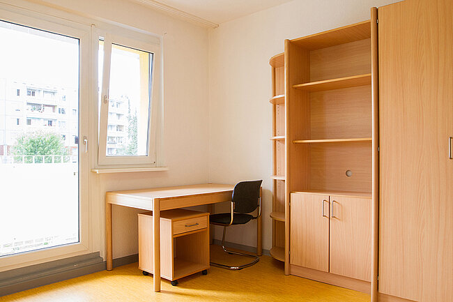 Ein einfaches Zimmer mit einem Schreibtisch, einem Stuhl und einem Regal. Der Schreibtisch steht vor einem großen Fenster, das Tageslicht hereinlässt. Das Regal bietet Stauraum und ist aus hellem Holz. Der Boden ist mit einem gelben Teppich bedeckt.