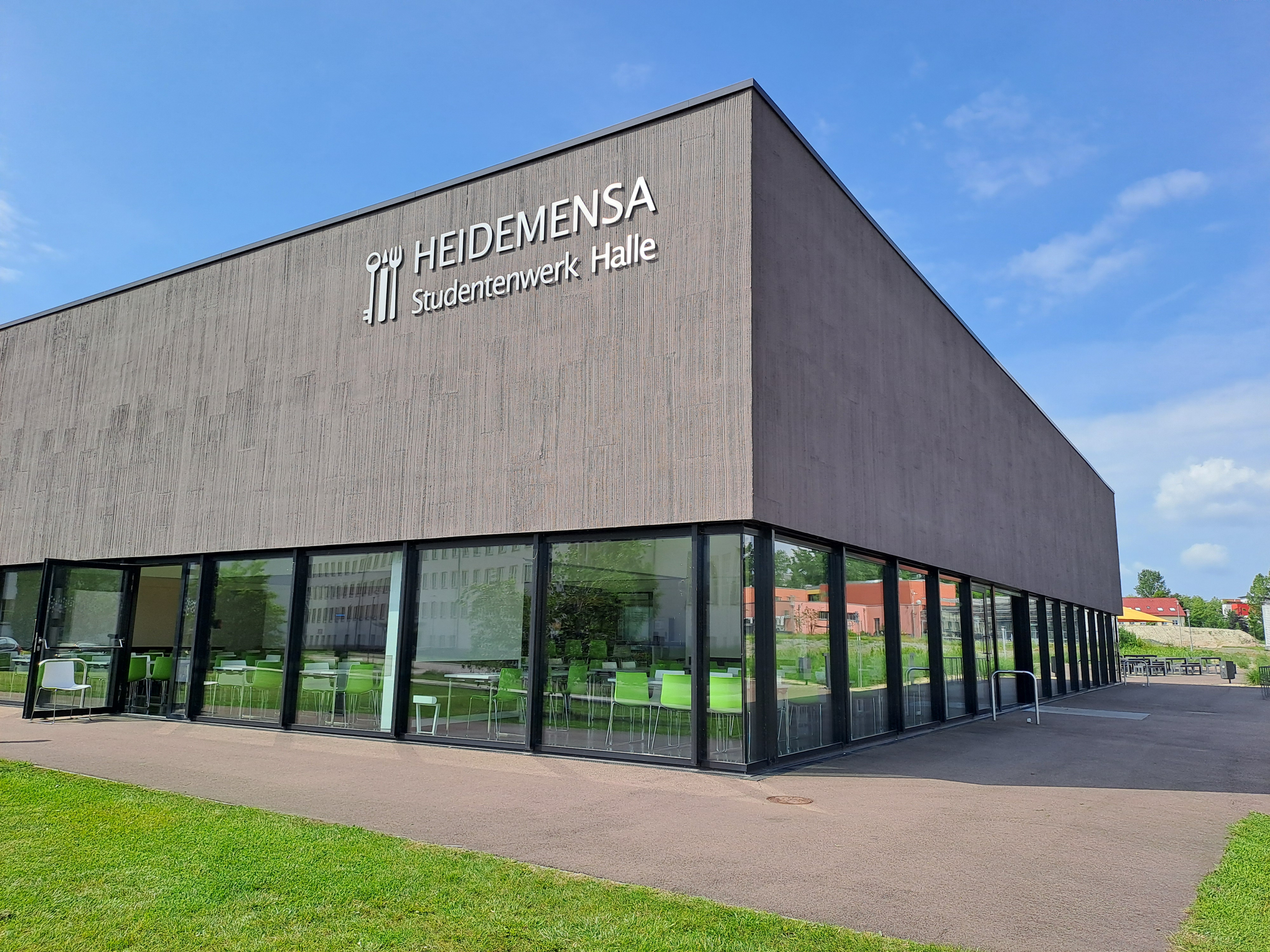 "Moderner Gebäudekomplex mit der Aufschrift 'HEIDEMENSA Studentenwerk Halle'. Das Gebäude hat eine graue Fassade mit großen Glasfronten und befindet sich auf einem grünen Gelände. Es handelt sich um eine Einrichtung des Studentenwerks in Halle."