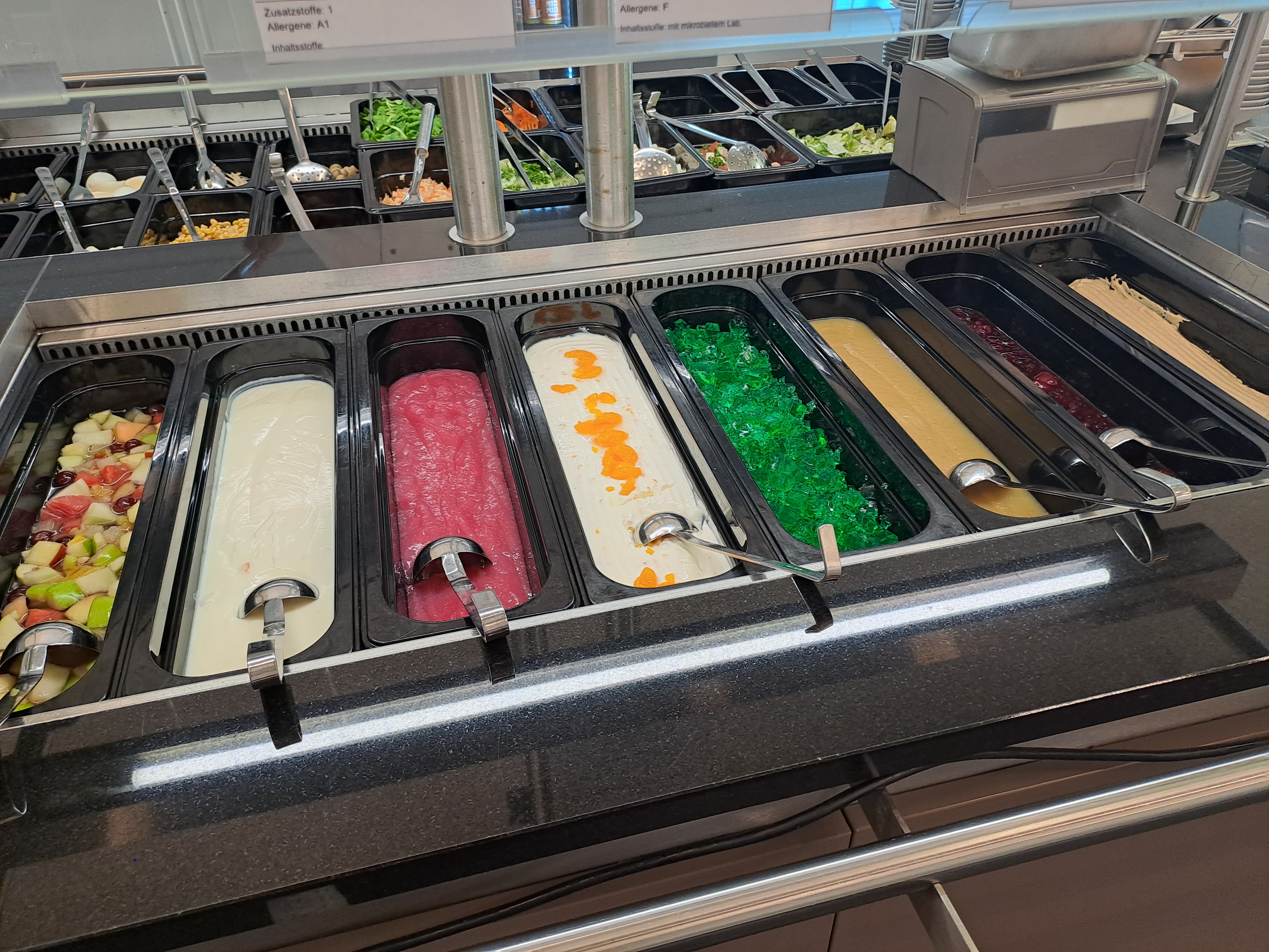 Ein Buffet mit verschiedenen Salatbar-Optionen. Es gibt verschiedene Salate, darunter einen grünen Salat, einen roten Salat, einen mit orangefarbenen Streifen und einen grünen Salat mit kleinen Brocken. Jeder Salat ist in separaten Behältern mit Servierlöffeln angeordnet.
