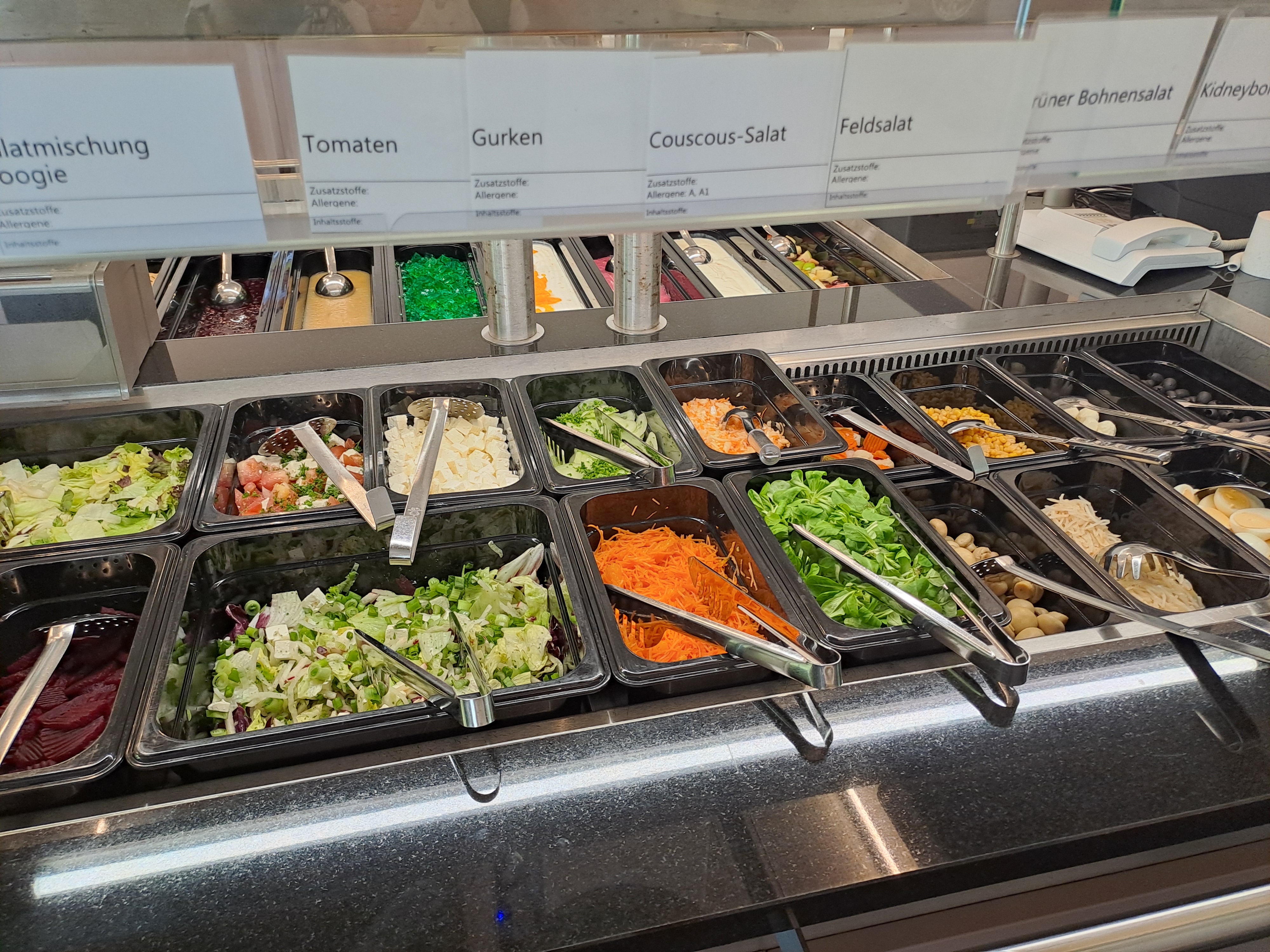 Ein Buffet mit verschiedenen Salaten. Von links nach rechts: Gemischter Salat, Tomaten, Gurken, Couscous-Salat, Feldsalat, grüne Bohnensalat und Kidneybohnen. Jede Schüssel ist mit einer Beschriftung versehen und enthält frische Zutaten.