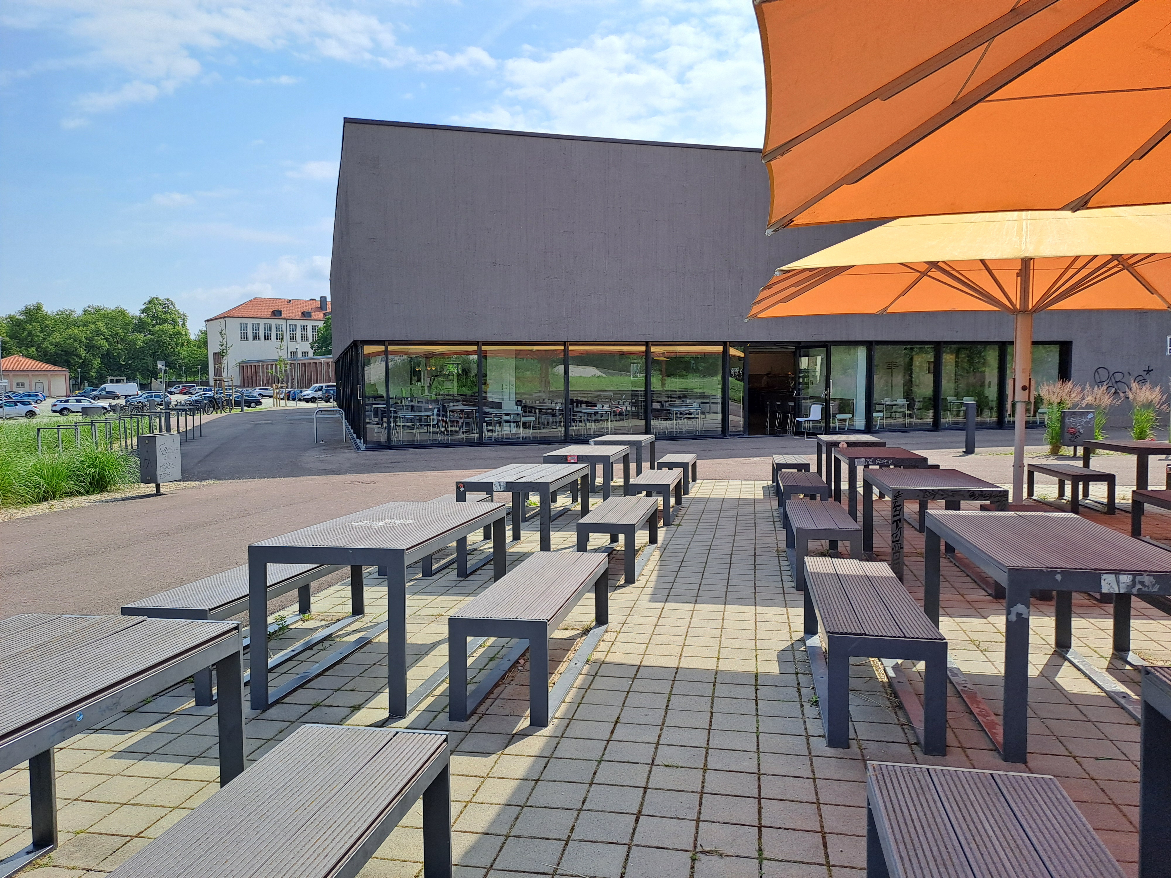 Ein Außenbereich mit mehreren Holzbänken und Tischen, die auf einem gepflasterten Boden aufgestellt sind. Große orange Sonnenschirme bieten Schatten. Im Hintergrund befindet sich ein modernes Gebäude mit großen Fenstern und einer überdachten Terrasse. Die Umgebung wirkt sauber und einladend.